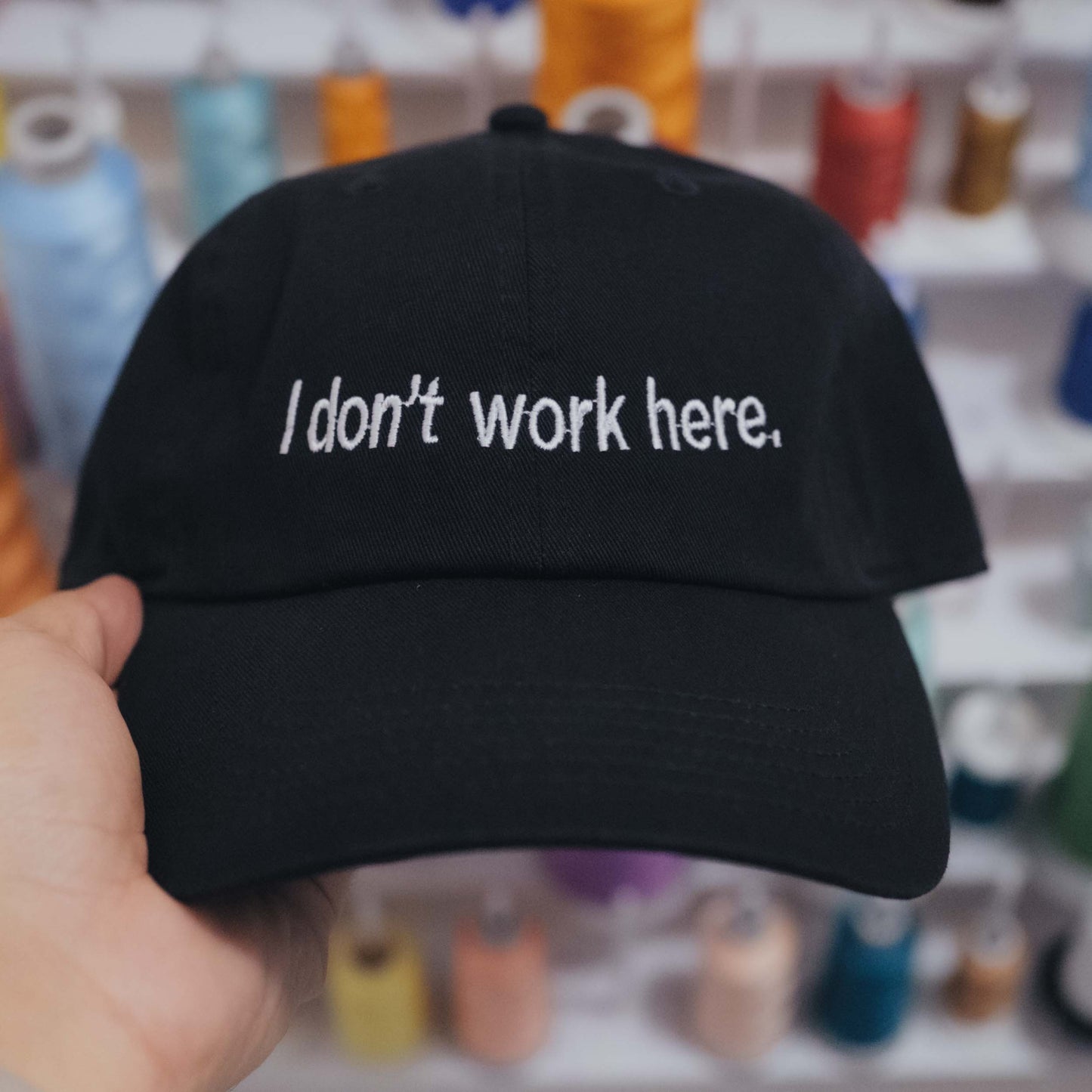 Black cap with 'I don't work here.' text held in front of colorful spools of thread.