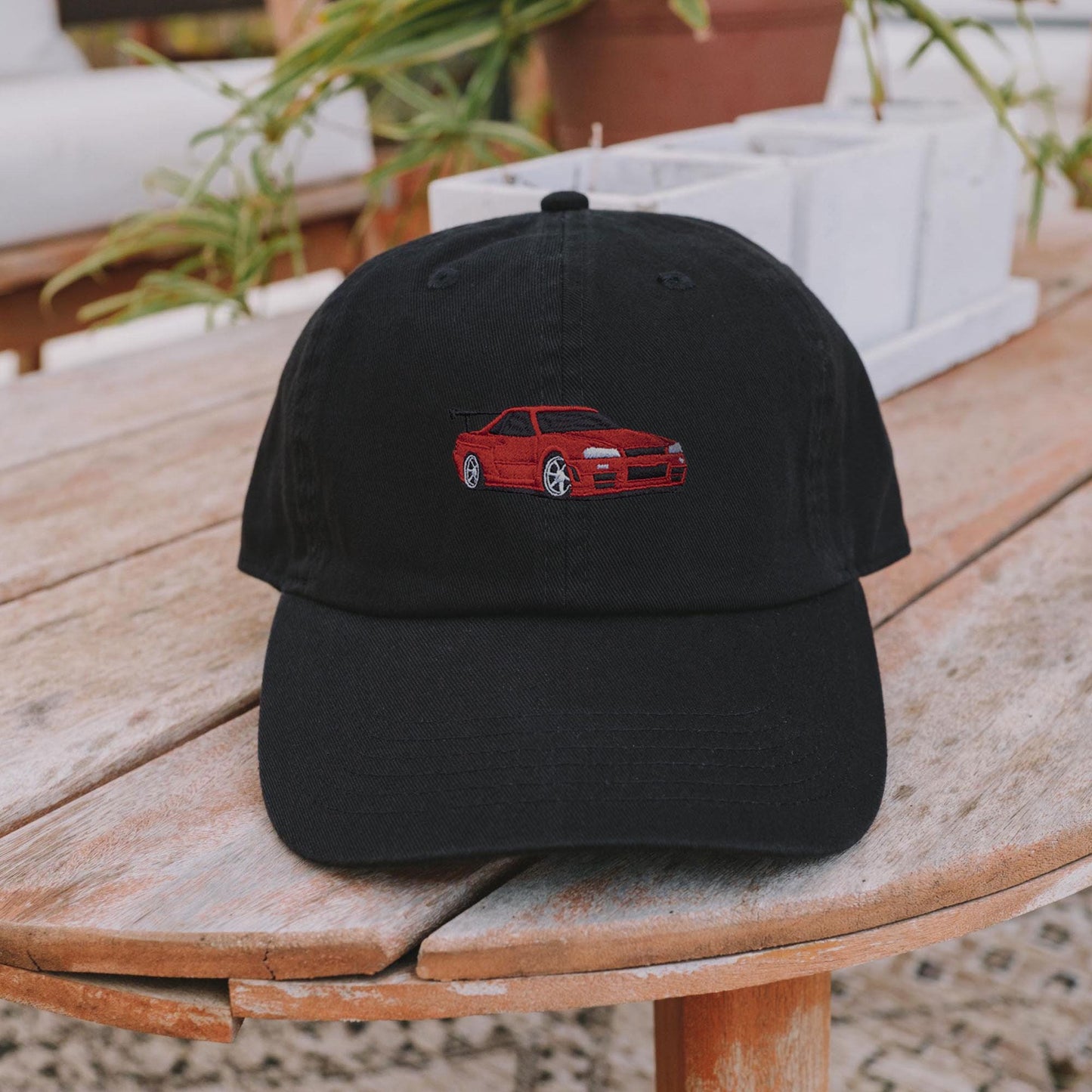 May include: A black baseball cap featuring a red embroidered car design. The car is a sports car with a spoiler. The cap has a curved brim and a button on top. The background includes plants and a wooden surface.