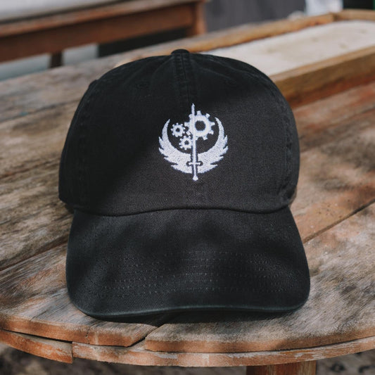 A black cotton dad hat with a white embroidered 'Brotherhood of Steel' symbol from the Fallout series.