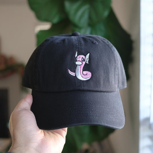 A black embroidered dad hat with a pink and white Dratini design on the front, held in a hand against a blurred background.