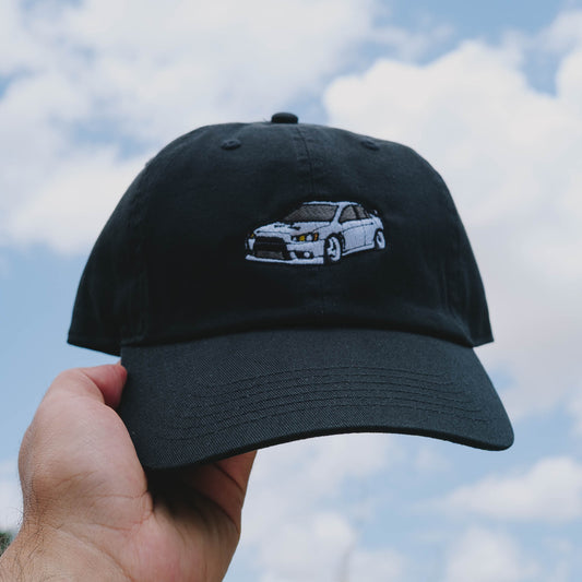 Black cap with embroidered car design held against a cloudy sky
