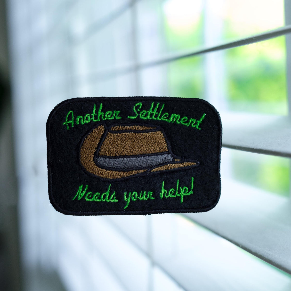 May include: Black rectangular patch with green embroidered text and a brown fedora design. The text reads "Another Settlement Needs your help!" The patch is likely for a jacket or bag.