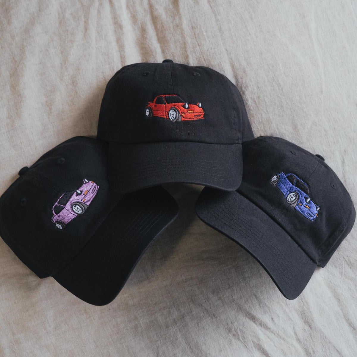May include: Three black baseball caps with embroidered images of cartoon cars. The cars are red, purple, and blue.