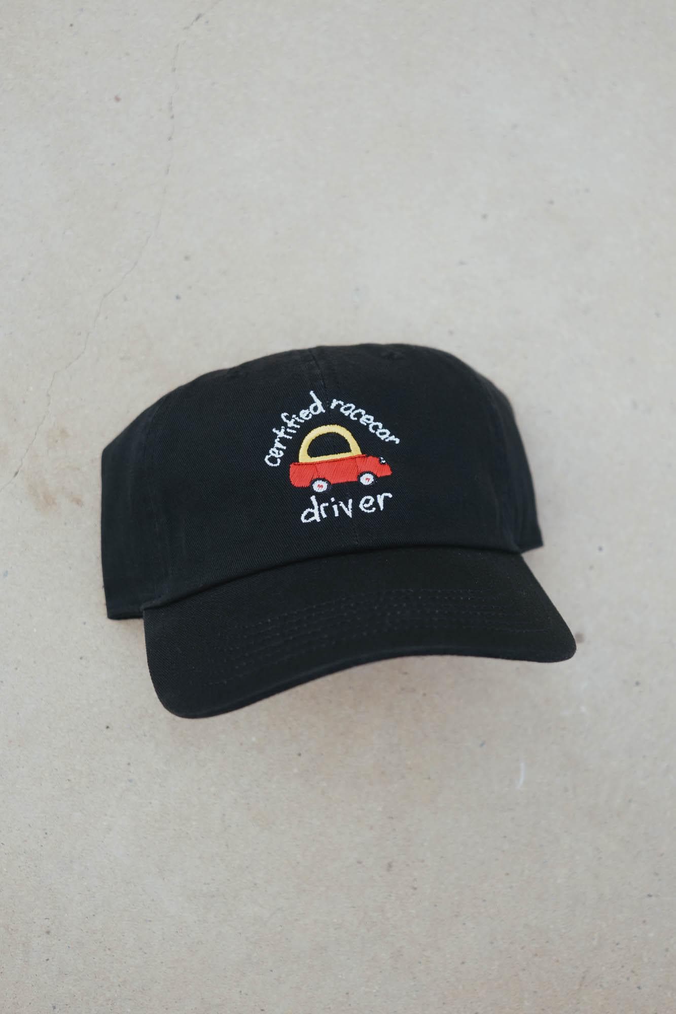 May include: Black baseball cap with a curved brim. The cap features an embroidered design of a red racecar with a yellow arch and the words "certified racecar driver" in white script. The cap is made of a soft fabric.