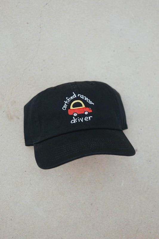 May include: Black baseball cap with a curved brim. The cap features an embroidered design of a red racecar with a yellow arch and the words "certified racecar driver" in white script. The cap is made of a soft fabric.