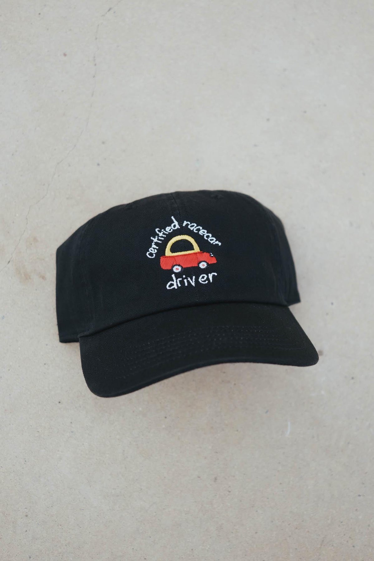 May include: Black baseball cap with a curved brim. The cap features an embroidered design of a red racecar with a yellow arch and the words "certified racecar driver" in white script. The cap is made of a soft fabric.
