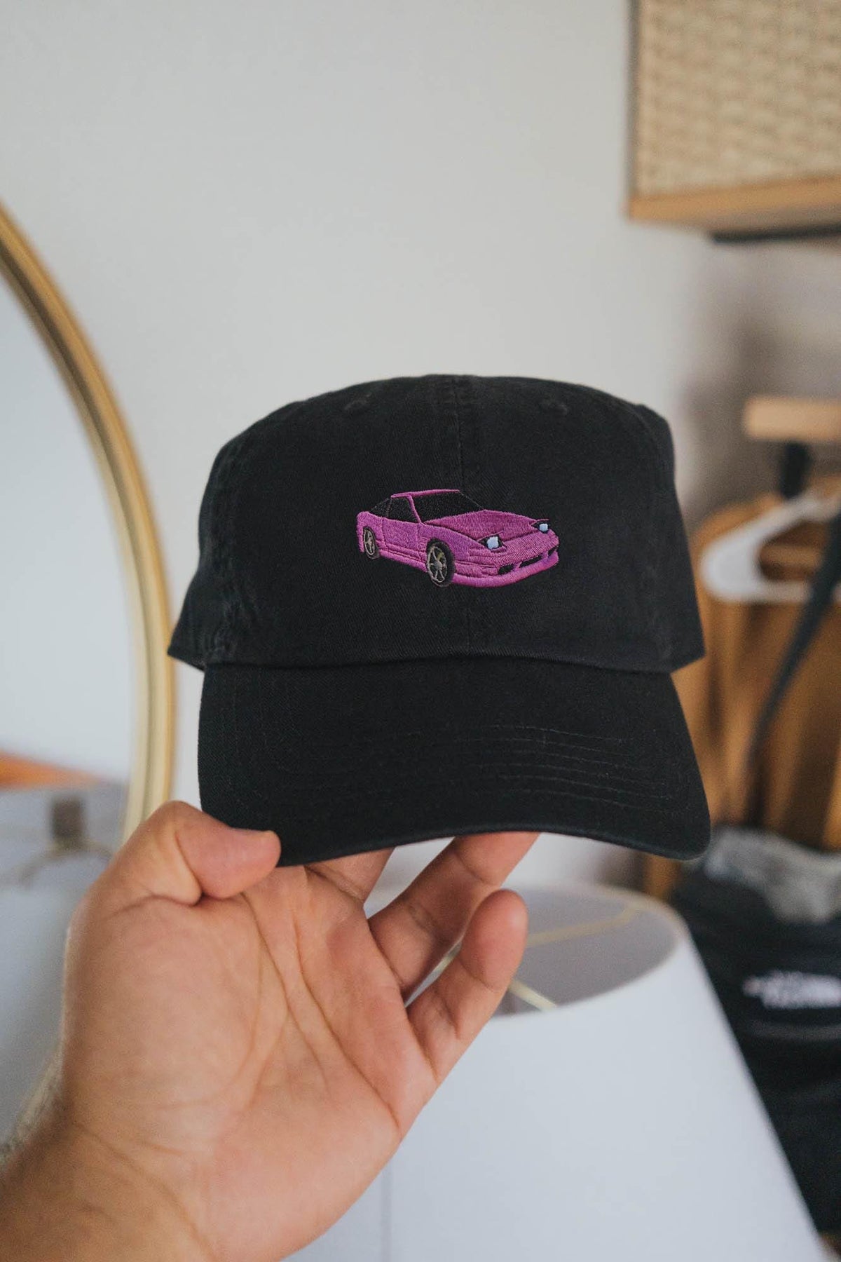 May include: Black baseball cap with a pink embroidered car design on the front. The cap has a curved brim and an adjustable strap at the back. The car design is a stylized representation of a sports car.