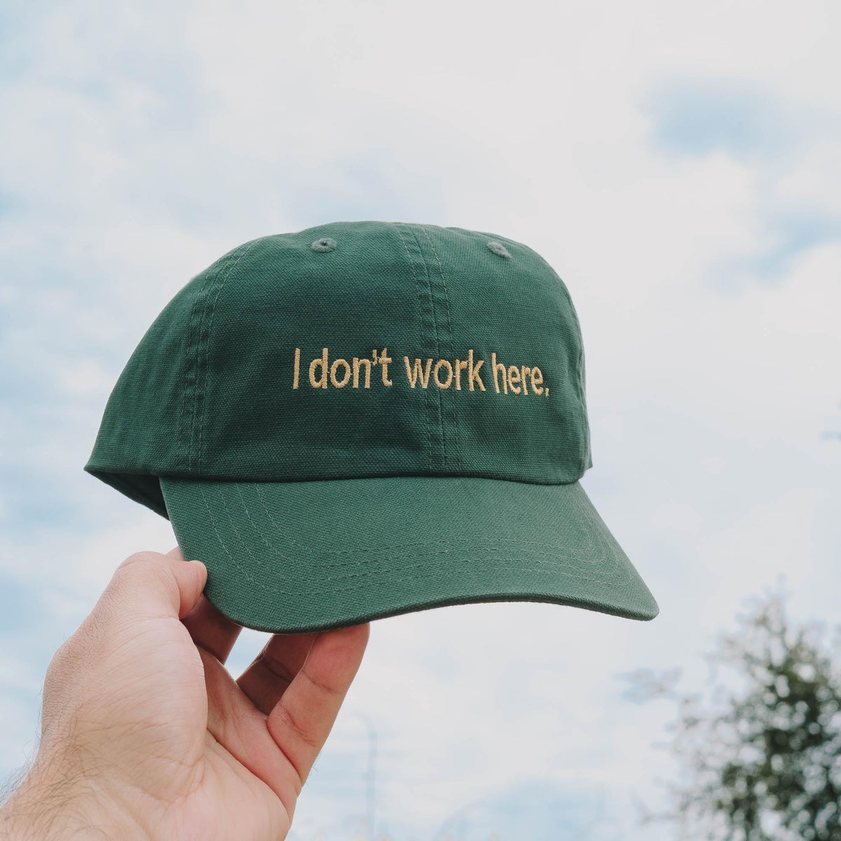 May include: A forest green baseball cap featuring the embroidered text I don't work here, in a gold-colored thread. The cap has a structured crown, a curved brim, and a button on the top center, showcasing a simple and functional design.