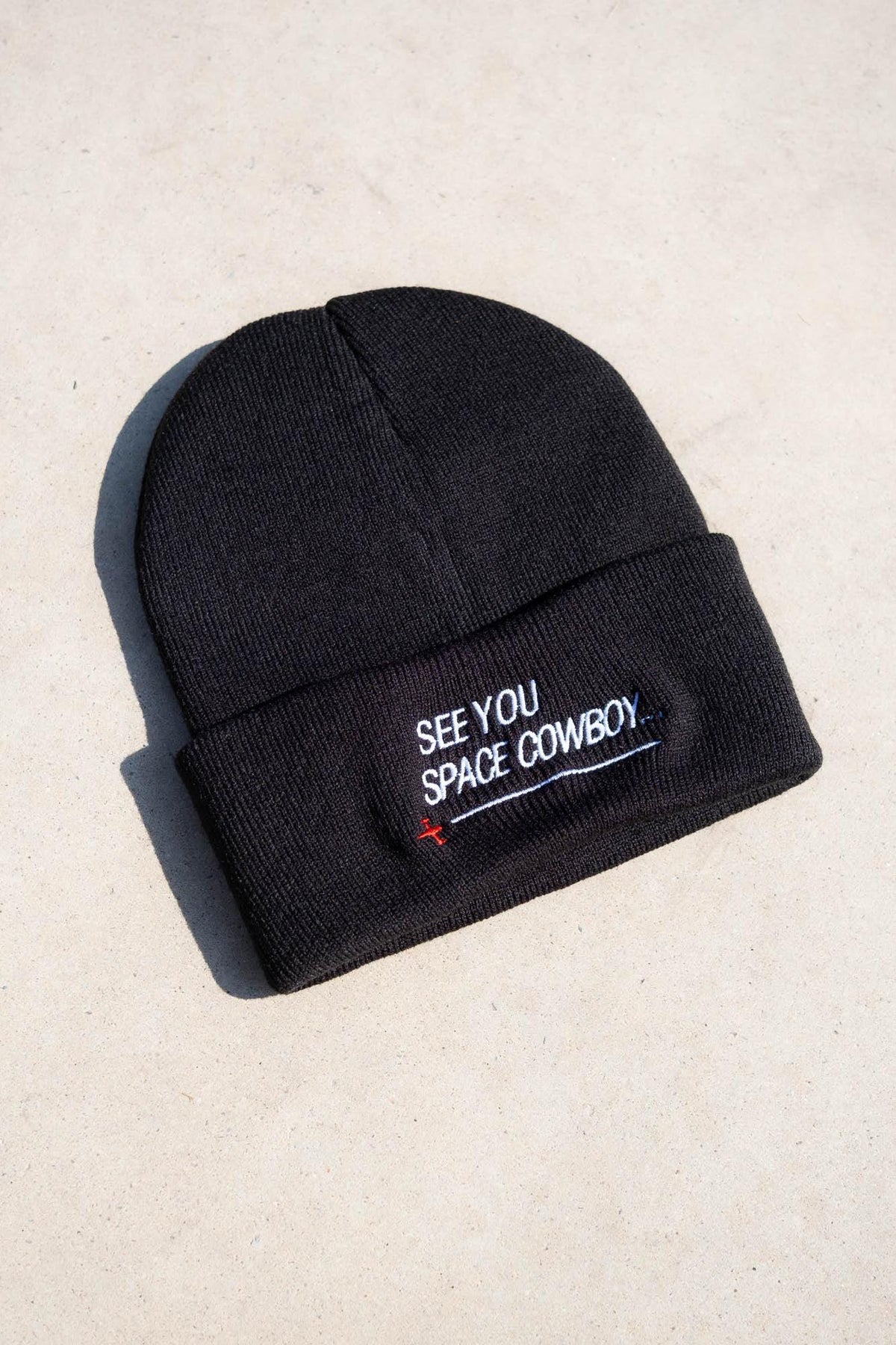 May include: Black knit beanie with a folded brim. The beanie features white embroidered text that reads "SEE YOU SPACE COWBOY" with a small red cross below the text. The hat is made of a soft, knitted material.